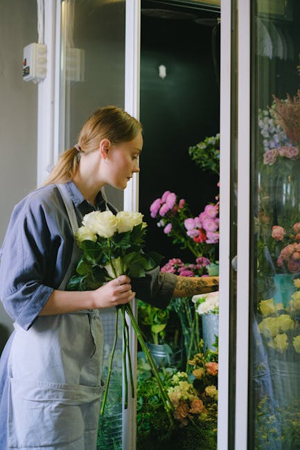 A young woman with auburn hair tied back, wearing a grey apron over a dark shirt, is selecting a bouquet of white roses with lush green stems and leaves at a flower shop. She holds the bouquet in both