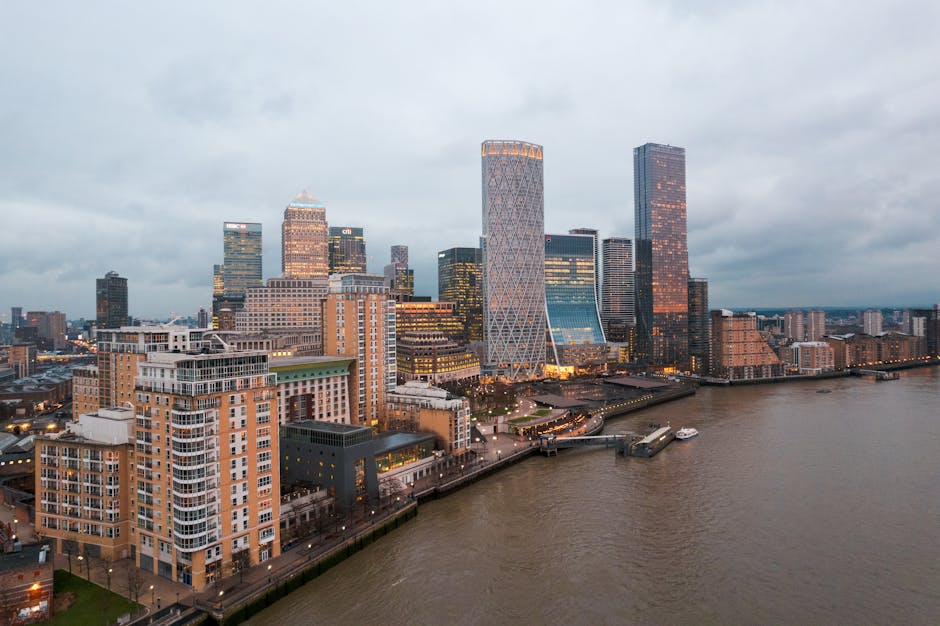 A panoramic view of Canary Wharf in London during early evening, showcasing a cluster of modern skyscrapers with glass facades reflecting the fading light. The tallest buildings include a distinctive