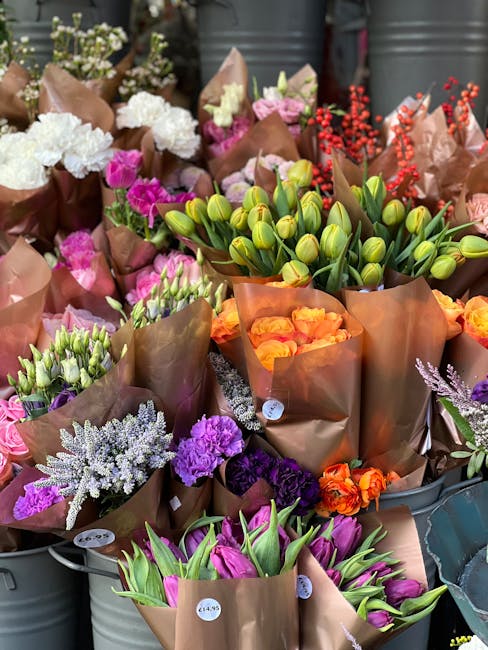 A vibrant display of fresh flowers arranged in brown kraft paper wrapping, featuring a variety of blooms including tulips in shades of purple and green, tulip buds, bright orange roses, purple and whi