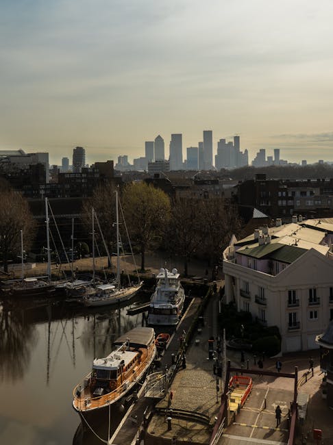A skyline view of Canary Wharf in London with high-rise office buildings under a partly cloudy sky, seen from a residential area with a small marina. In the foreground, a boat is docked alongside a wa