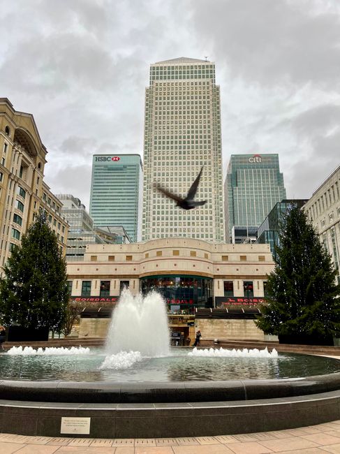 A cityscape view showing the HSBC and Citi bank skyscrapers behind a large, ornate building with a curved facade, situated in a busy urban square. In the foreground, there is a fountain with water spl