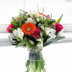 Combining architectural structure and organic beauty, this striking bouquet captures the essence of Canary Wharf's unique blend of modernity and riverside tranquility. Towering above a clear, slender vase, the arrangement is crowned with bold orange and creamy-peach gerberas, their voluminous petals radiating warmth and a gentle optimism. Rich fuchsia roses lend luxuriant vibrancy, their tightly coiled centres hinting at deeper feelings, ideally suited for celebrations or thoughtful gestures around Westferry or the Isle of Dogs. Pristine white alstroemeria and spray roses cluster gracefully among the larger blooms, scattering points of light that recall reflections on the docks at sunset. Soft hints of blush pink and whimsical filler flowers build out the bouquet's lush profile, nestled among an abundance of deep, polished foliage. Aspidistra leaves, wide and sturdy, curve around the base in a protective, artful flourish, while smaller, pointed greens weave a sense of movement throughout. Illuminated by a soft, contemporary background, the entire arrangement exudes freshness, vitality, and a sense of occasion-making it a beautiful expression for celebrations, anniversaries, or simply adding a personal touch to a Canary Wharf office or penthouse.